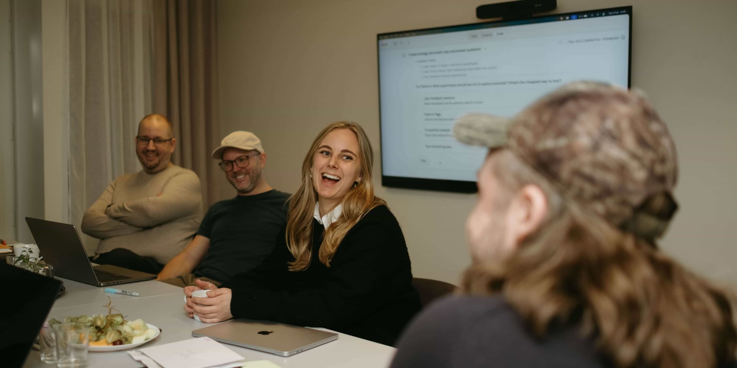 Four of the team laughing around the meeting-room table, a laptop open and a screen on the wall behind them